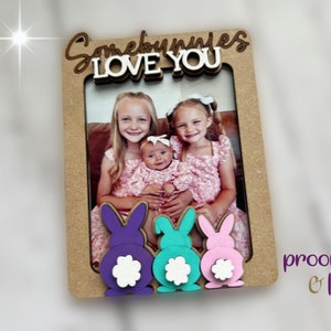Peut inclure: Un cadre photo en bois avec les mots "Somebunnies LOVE YOU" en &eacute;criture cursive. Le cadre pr&eacute;sente trois queues de lapin color&eacute;es en violet, turquoise et rose. Le cadre contient une photo de trois jeunes filles.