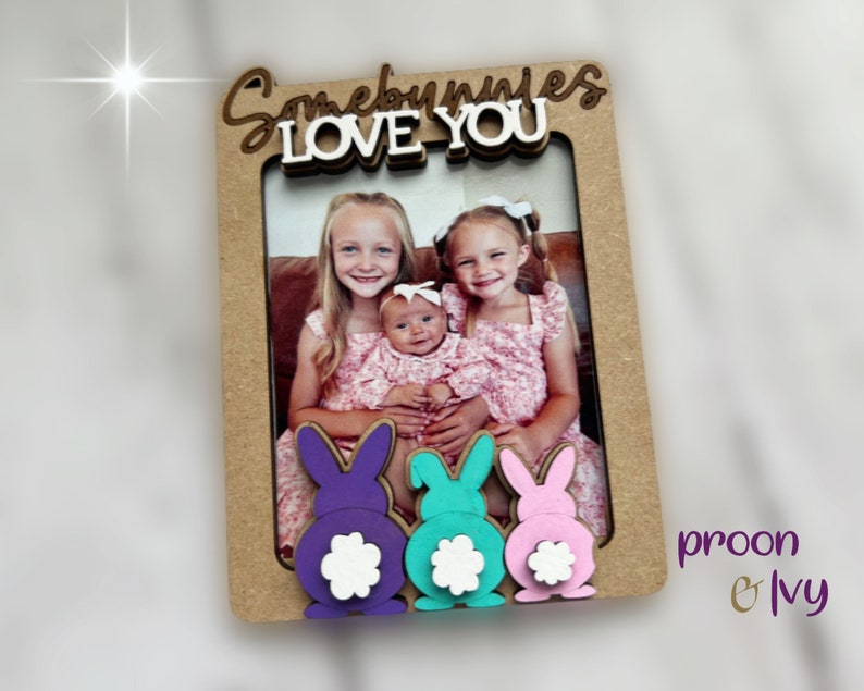 Peut inclure: Un cadre photo en bois avec les mots "Somebunnies LOVE YOU" en &eacute;criture cursive. Le cadre pr&eacute;sente trois queues de lapin color&eacute;es en violet, turquoise et rose. Le cadre contient une photo de trois jeunes filles.