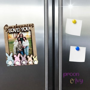 Peut inclure: Un cadre photo en bois avec les mots "Somebunnies LOVE YOU" et des d&eacute;coupes de lapins. Le cadre contient une photo de famille et est fix&eacute; &agrave; un r&eacute;frig&eacute;rateur avec des aimants. Les lapins sont de couleurs pastel.