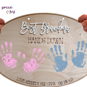 May include: A wooden oval sign with the text "Best Grandma Hands Down" in black lettering. The sign also features two pink handprints and two blue handprints, along with the text "Love Kennedy and Capri 05-14-23".