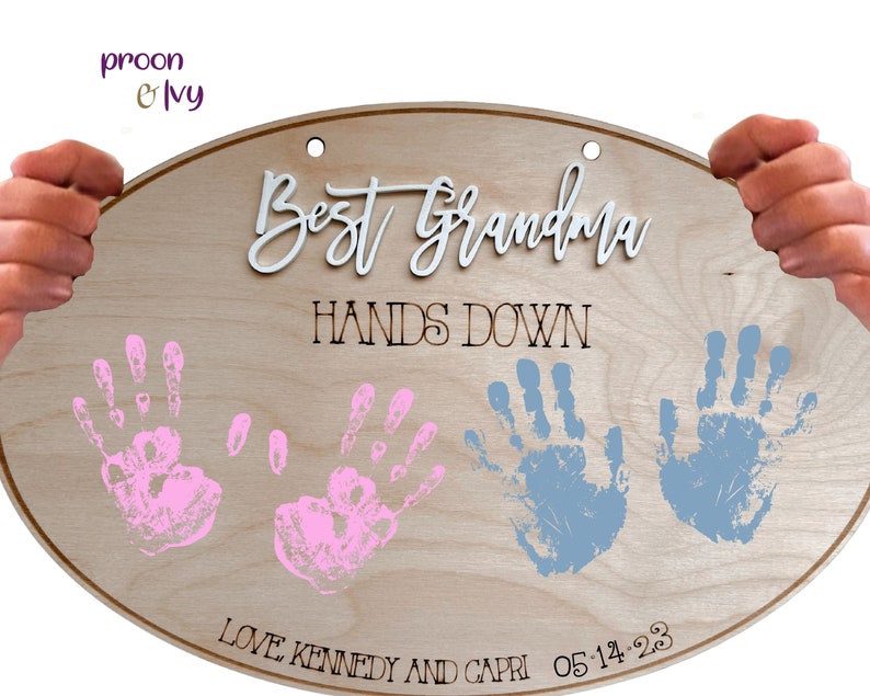May include: A wooden oval sign with the text "Best Grandma Hands Down" in black lettering. The sign also features two pink handprints and two blue handprints, along with the text "Love Kennedy and Capri 05-14-23".
