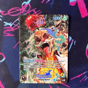 May include: A trading card featuring a colorful illustration of a character named Vegapunk. The card has a vibrant design with text and numbers, including "5000" and "2". The card is from the "Scientist / Egghead" series.
