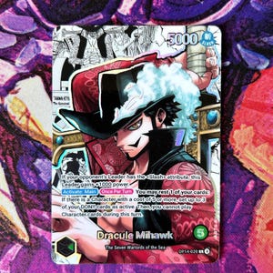 May include: A One Piece trading card featuring Dracule Mihawk. The card has a detailed illustration of the character with a black hat and a red and black outfit. The card includes text describing the character's abilities and a power level of 5000.