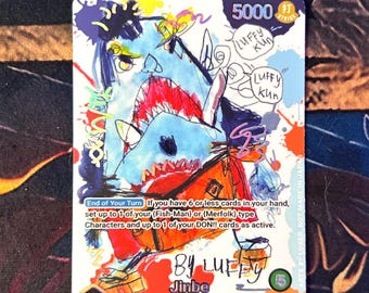 以下が含まれることがあります： 「BY LUFFY Jinbe Fish-man/Strat Hat Crew」のテキストが入った、サメのような外観のキャラクターのカラフルなイラストが描かれたトレーディングカード。 カードには5000のストライク値と、ターンの終了に関するテキストが含まれています。