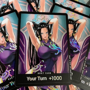 May include: A stack of trading cards featuring a woman in a purple dress with long black hair. The cards have the text "DON!! CARD" and "Your Turn +1000". The background has a light blue and pink heart design.