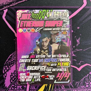 Può includere: Una carta collezionabile rettangolare con sfondo rosa e viola. La carta presenta un'illustrazione di un personaggio con i capelli blu e il testo "Jinx Etherium Shaper Legendary Artifact Creature". La carta include anche le istruzioni di gioco e la firma dell'artista.