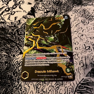 May include: A trading card featuring Dracule Mihawk from "The Seven Warlords of the Sea." The card has a green and gold design with the number 5000 in the upper right corner. The card text describes the character's abilities. The card is marked OP14-020.
