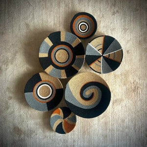 Könnte beinhalten: Sechs gewebte Wanddekoration Körbe in verschiedenen Größen und Mustern. Die Körbe sind aus natürlichen Materialien gefertigt und weisen geometrische Muster in Grautönen, Schwarz und Braun auf.