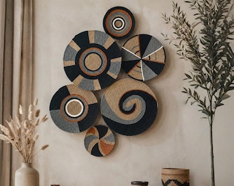 May include: A collection of six circular wall-mounted baskets arranged in a cluster. The baskets feature coiled designs in shades of black, gray, tan, and orange, with geometric patterns, concentric circles, and spiral motifs throughout the set.