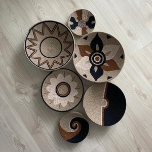 May include: Six woven wall baskets with geometric patterns in black, brown, and white. The baskets are arranged in a circular pattern on a light wood floor.