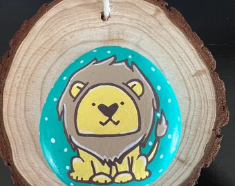 May include: A wooden slice with a painted rock featuring a cartoon lion with brown mane, yellow body, and white spots on a teal background.