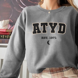 Puede incluir: Sudadera gris con el texto "ATYD EST. 1971" y un gráfico de luna creciente.