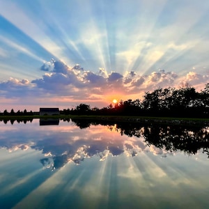 May include: A scenic view of a lake reflecting a vibrant sunset sky with streaks of light. The sky is a mix of blue, pink, and orange with fluffy white clouds. The lake is calm and still, reflecting the colors of the sky.