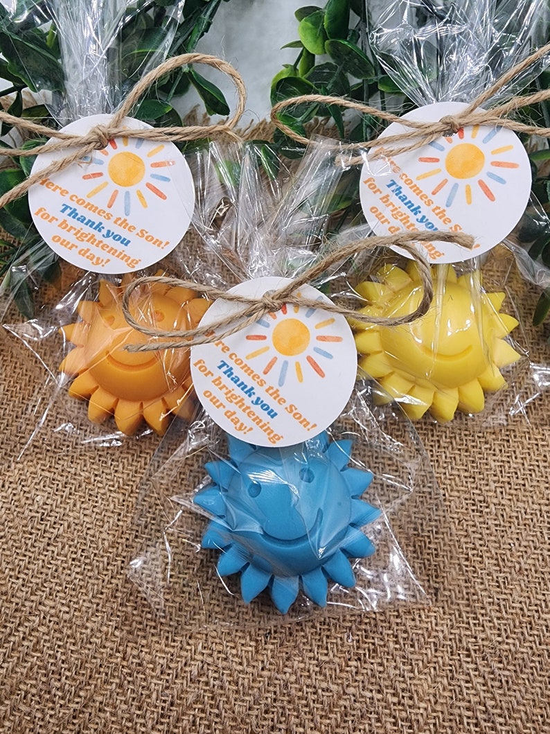 May include: Three small sun-shaped favours wrapped in clear cellophane bags with twine bows. Each favour has a round tag attached with the text "Here comes the Son! Thank you for brightening our day!"