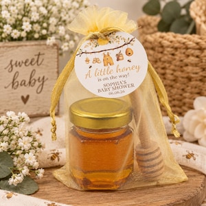 May include: A small hexagonal glass jar filled with golden honey, featuring a gold-colored lid, placed inside a sheer yellow organza drawstring bag with a wooden honey dipper and a round tag printed with A little honey is on the way!