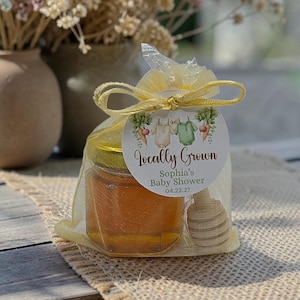 May include: A small glass jar filled with amber honey, enclosed in a yellow organza drawstring bag with a wooden honey dipper. A round white tag is tied to the bag, featuring illustrations of baby onesies and the text Locally Grown, Sophia's Baby Shower, 04.22.27.