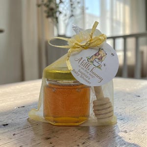 May include: A small glass jar filled with golden honey, featuring a gold lid and a wooden honey dipper, enclosed in a yellow organza drawstring bag. A round white tag is tied to the top, displaying a Winnie the Pooh illustration and the text A little hunny is on the way! Sophia's Baby Shower.