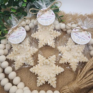 May include: Snowflake-shaped soaps in clear cellophane bags, tied with twine and a circular tag that reads "Happy Holidays From The Williams 2025." The soaps are off-white and sit on a burlap surface.
