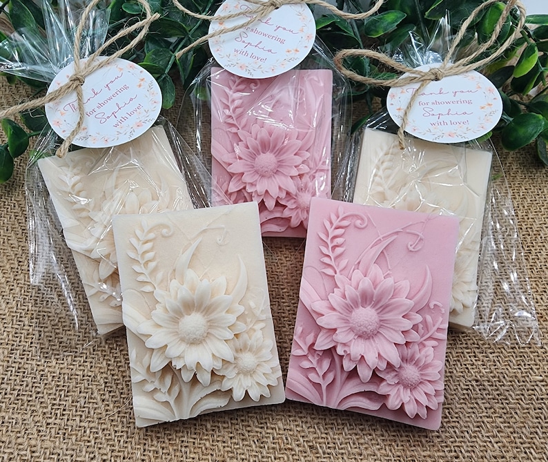 Puede incluir: Cuatro peque&ntilde;as barras de jab&oacute;n rectangulares envueltas en celof&aacute;n transparente. Dos barras son blancas con un dise&ntilde;o floral y dos barras son rosas con un dise&ntilde;o floral. Cada barra tiene una peque&ntilde;a etiqueta redonda adjunta con el texto "&iexcl;Gracias por duchar a Sophia con amor!"
