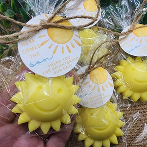 May include: Yellow sun-shaped soap favours with a smiling face, packaged in clear cellophane bags with a tied twine bow. Each bag includes a round tag with the text "Here comes the son" and "Thank you for brightening our day!"
