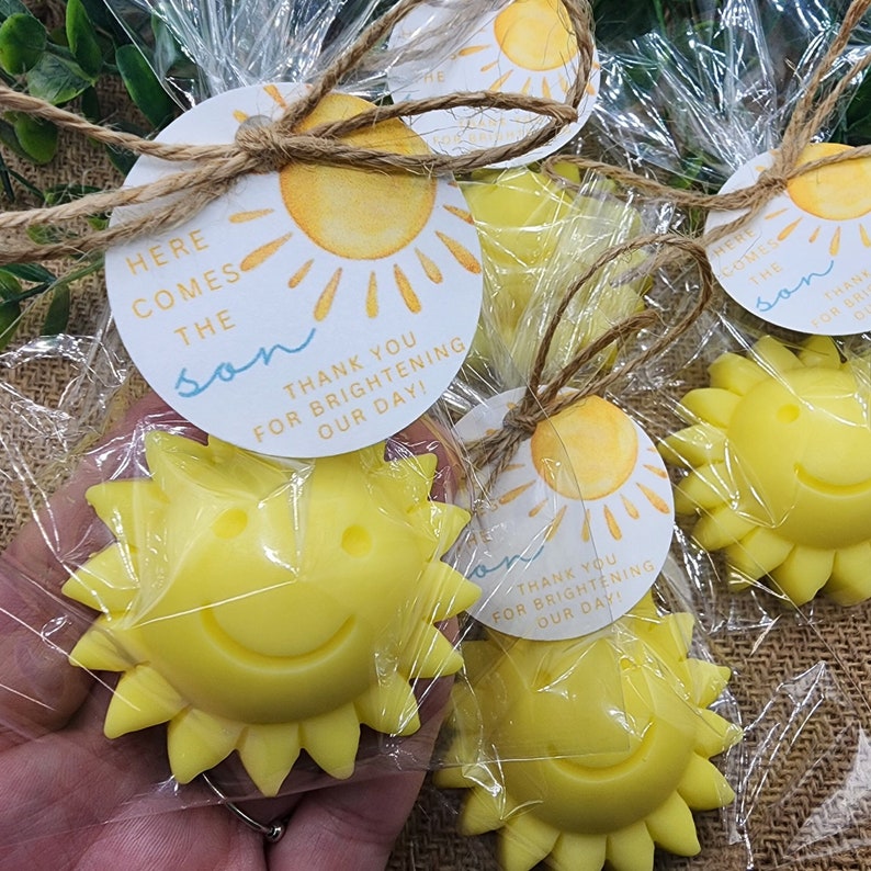 May include: Yellow sun-shaped soap favours with a smiling face, packaged in clear cellophane bags with a tied twine bow. Each bag includes a round tag with the text "Here comes the son" and "Thank you for brightening our day!"