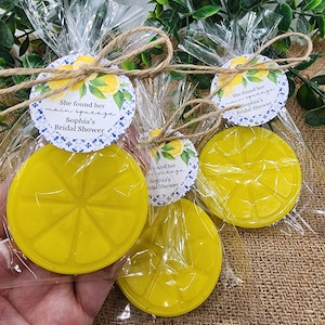 May include: Three clear plastic bags with yellow lemon-shaped soap bars and a round tag tied with twine. The tag has a blue and white patterned background with lemons and the text "She found her main squeeze Sophia's Bridal Shower".