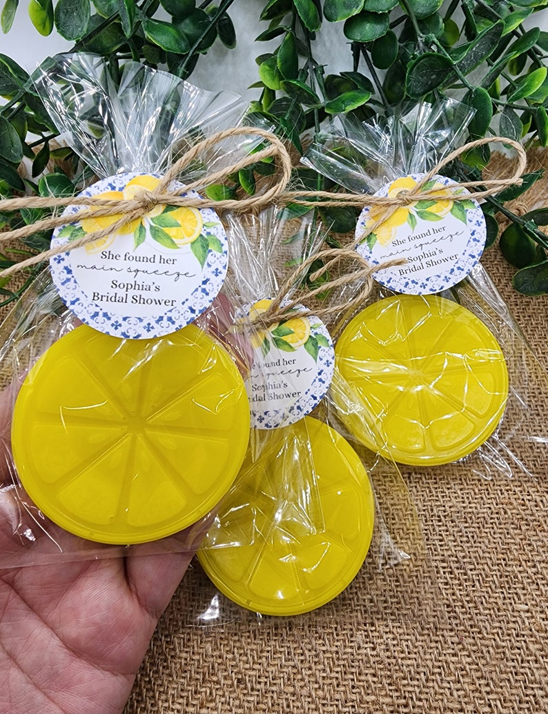 May include: Three clear plastic bags with yellow lemon-shaped soap bars and a round tag tied with twine. The tag has a blue and white patterned background with lemons and the text "She found her main squeeze Sophia's Bridal Shower".