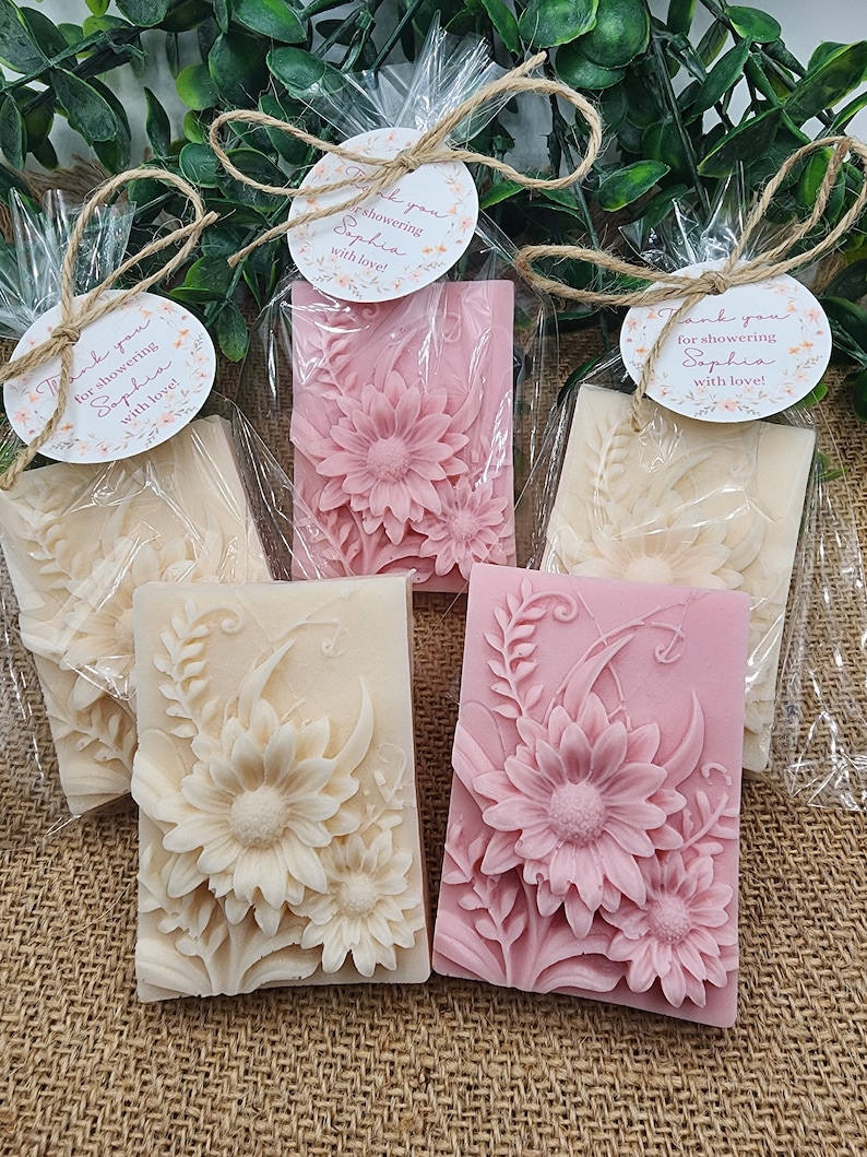 Puede incluir: Cuatro peque&ntilde;as barras rectangulares de jab&oacute;n con dise&ntilde;os florales. Dos barras son rosas con flores blancas y dos barras son blancas con flores rosas. Cada barra est&aacute; envuelta en celof&aacute;n transparente y atada con una cuerda. Una peque&ntilde;a etiqueta redonda con el texto "&iexcl;Gracias por duchar a Sophia con amor!" est&aacute; unida a cada barra.