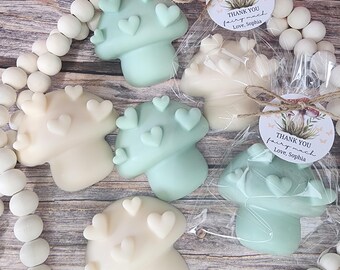 May include: A collection of mushroom-shaped soaps in shades of white and mint green, each adorned with heart-shaped accents. Some are packaged with a tag that reads "THANK YOU fairy much Love, Sophia". The soaps are arranged on a wooden surface.