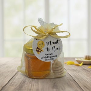 May include: A small glass jar filled with golden honey, enclosed in a sheer yellow organza drawstring bag with a wooden honey dipper. A round white tag features a cartoon bee and the text: Meant to Bee! Anna's Bridal Shower 03.20.2027.