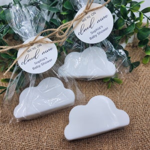 May include: Three white cloud-shaped soaps in clear cellophane bags, tied with twine and a round tag. The tag reads "We're on cloud nine Sophia's Baby Shower". The soaps are on a burlap surface with greenery.