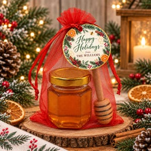 May include: A small hexagonal glass jar filled with golden honey, featuring a gold lid and a wooden honey dipper, enclosed in a red organza drawstring bag. A round tag attached to the bag reads Happy Holidays from THE WILLIAMS with botanical art.
