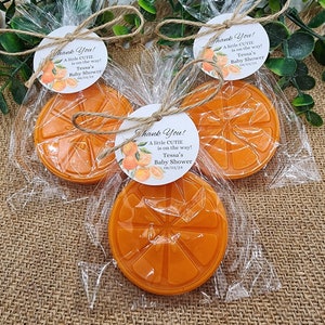 May include: Three orange-shaped soaps wrapped in clear cellophane bags with a white tag tied with twine. The tag has a floral design and the text "Thank You! A little CUTIE is on the way! Tessa's Baby Shower 06/01/24"
