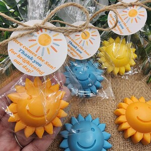 May include: A set of four small, colourful, smiling sun-shaped soaps wrapped in clear cellophane bags with a brown twine bow and a round tag that says "Here comes the Son! Thank you for brightening our day!"