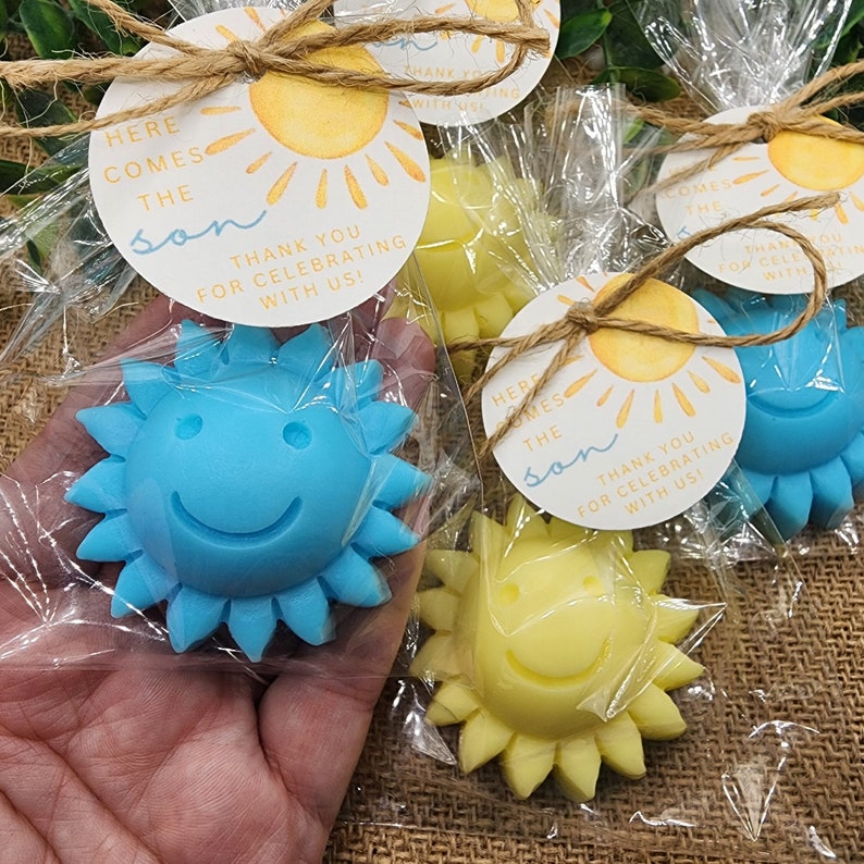 May include: A set of favour bags for a baby shower. Each bag contains a small yellow or blue sun-shaped soap with a smiling face. The bags are tied with twine and have a tag that says "Here comes the son. Thank you for celebrating with us!"