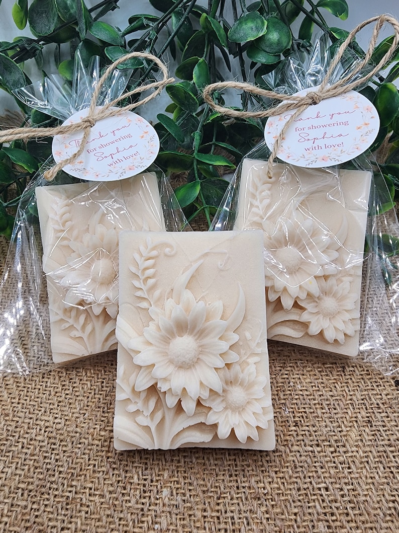 Puede incluir: Tres peque&ntilde;os jabones blancos con forma de flores envueltos en bolsas de celof&aacute;n transparentes con un lazo de cuerda marr&oacute;n y una peque&ntilde;a etiqueta redonda blanca con texto rosa que dice "&iexcl;Gracias por duchar a Sophia con amor!"