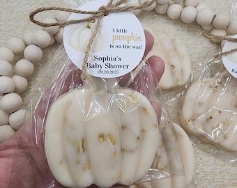 May include: Pumpkin-shaped soap favors for a baby shower. The soaps are white with gold flecks and are packaged in clear cellophane bags. Each favor has a round tag with the text "A little pumpkin is on the way!" and "Sophia's Baby Shower 09.20.2025".
