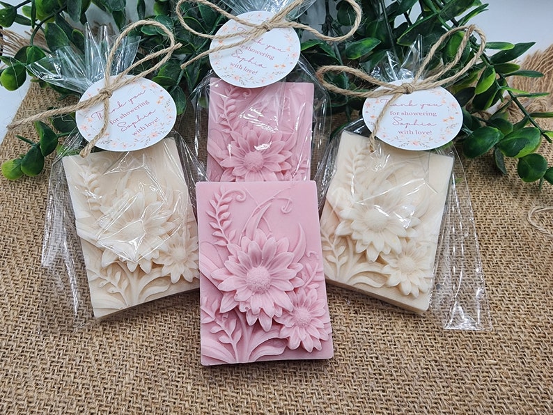 Puede incluir: Tres peque&ntilde;as bolsas de regalo con envoltura de celof&aacute;n transparente, cada una contiene una pastilla de jab&oacute;n con dise&ntilde;o floral. Las pastillas de jab&oacute;n son blancas y rosas con un dise&ntilde;o floral en relieve. Las bolsas est&aacute;n atadas con cuerda y tienen peque&ntilde;as etiquetas redondas con el texto "&iexcl;Gracias por celebrar a Sophia con amor!"