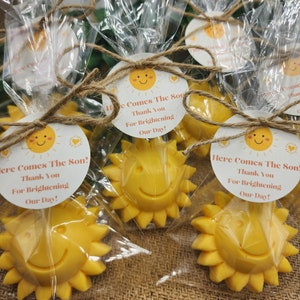 May include: A set of small yellow sun-shaped favours with a smiling face. Each favour is wrapped in clear cellophane and tied with twine. A round tag with the text "Here Comes The Son! Thank You For Brightening Our Day!" is attached to each favour.