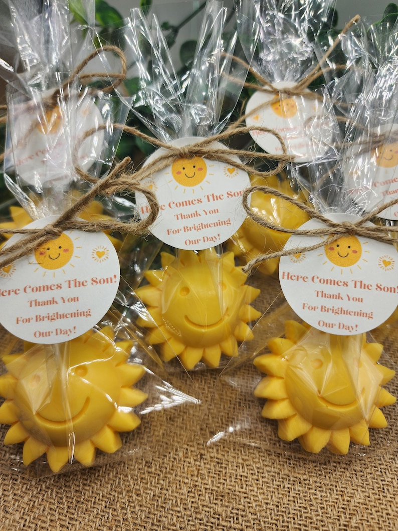 May include: A set of small yellow sun-shaped favours with a smiling face. Each favour is wrapped in clear cellophane and tied with twine. A round tag with the text "Here Comes The Son! Thank You For Brightening Our Day!" is attached to each favour.