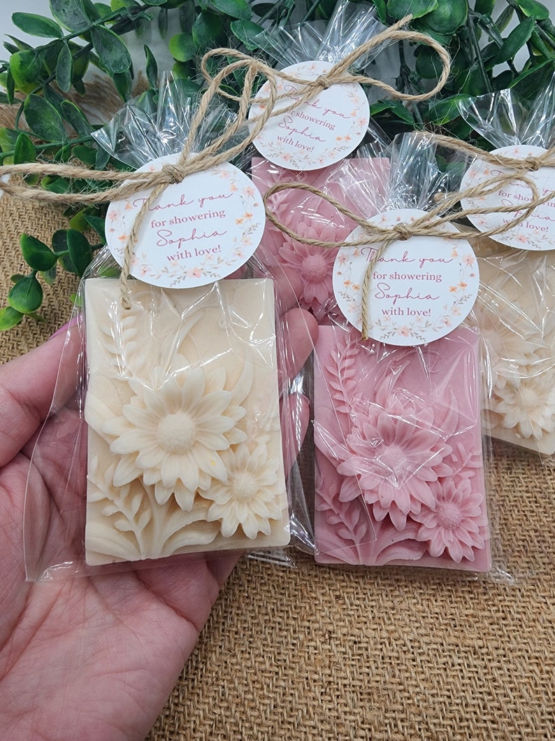 Puede incluir: Tres peque&ntilde;as barras de jab&oacute;n envueltas con dise&ntilde;os florales. Cada barra tiene una etiqueta redonda adjunta con el texto "&iexcl;Gracias por celebrar a Sophia con amor!" en letras rosas.