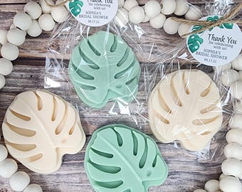 May include: Four leaf-shaped soaps in shades of green and cream, packaged in clear cellophane bags. Each soap has a monstera leaf design. The soaps are labeled "Sophia's Bridal Shower 09.17.25". Decorative wooden beads and twine are also visible.