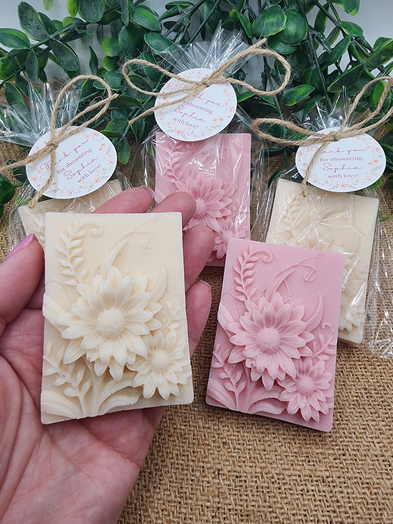 Puede incluir: Cuatro peque&ntilde;as barras rectangulares de jab&oacute;n con dise&ntilde;os florales. El jab&oacute;n est&aacute; envuelto en celof&aacute;n transparente y atado con cuerda. Cada jab&oacute;n tiene una peque&ntilde;a etiqueta redonda adjunta con el texto "&iexcl;Gracias por celebrar a Sophia con amor!"