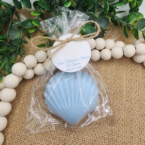 May include: A light blue seashell-shaped soap is packaged in clear cellophane and tied with twine. A round tag reads "From Miranda's Shower To Yours, March 7, 2026". The soap is surrounded by decorative elements.