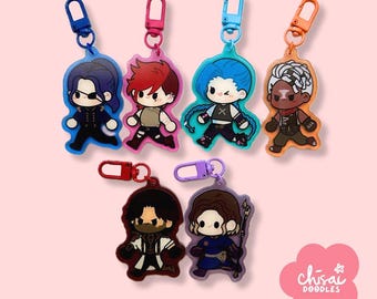 May include: A collection of colorful character keychains with various designs. Each keychain features a unique character with distinct features and outfits, attached to a metal clasp. The keychains are in shades of blue, red, orange, and purple, with a pink background.