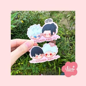May include: Two die-cut stickers featuring cartoon characters with the text "DOOMED YAOI". One sticker has two characters with light blue hair and the other has two characters with black hair. The stickers are held in front of a green leafy background.