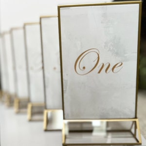 May include: Gold table number holder with a white card that says "One" in gold script.