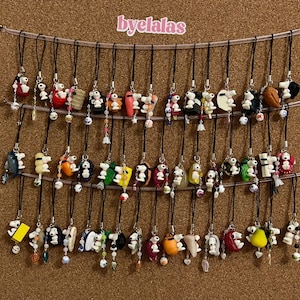 Peut inclure: Une collection de breloques Snoopy faites à la main, chacune avec un design et une palette de couleurs uniques. Les breloques sont suspendues à des cordons noirs et ornées de perles. Le fond est un tableau en liège, et le texte "byelalas" est visible.
