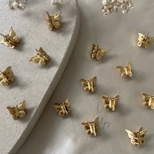 May include: A set of 12 gold butterfly hair clips. The clips are arranged on a white surface with a light brown background. The clips are all the same size and shape, and they are made of metal.