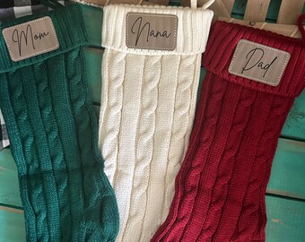May include: Three cable-knit Christmas stockings in green, cream, and red. Each stocking has a leatherette tag with the names "Mom", "Nana", and "Dad". The stockings have a loop for hanging.